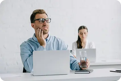 Vīrietis ar brillēm birojā turas pie vaiga un žokļa, ar otru roku turot ūdens glāzi, attēlojot akūtas zobu sāpes