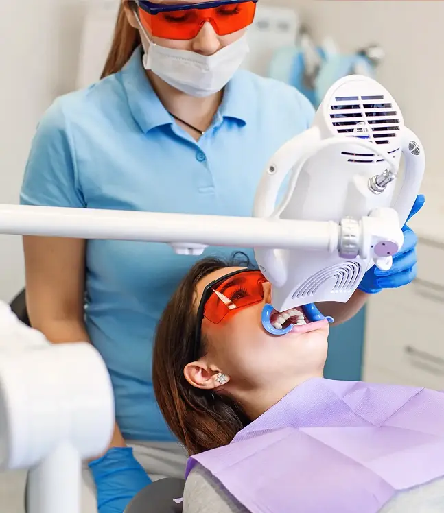 Paciente zobārstniecības krēslā, tiek veikta zobu balināšanas procedūra ar lampu un aizsargbrillēm